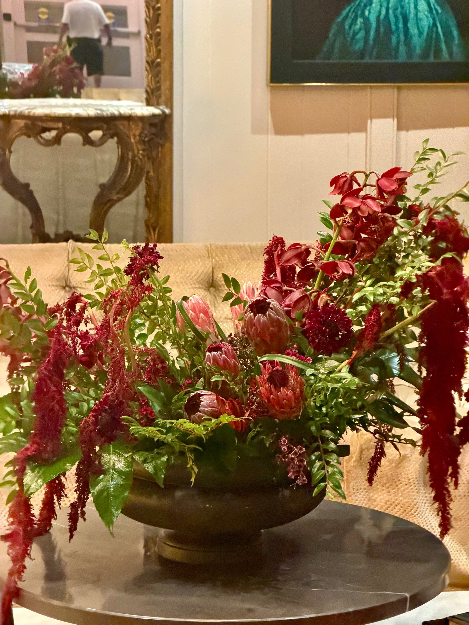 A large, lush Charleston hotel floral arrangement in a dark bronze urn-shaped bowl, set on a round marble table in front of a beige tufted sofa. The arrangement features deep red amaryllis, red fuchsia, green foliage, and a central protea, with a warm, elegant interior background.